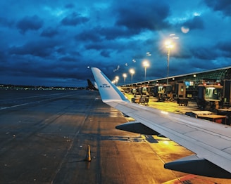 An airport runway is visible during dusk, with the wing of a KLM airplane in the foreground. Bright lights illuminate the tarmac and terminal, contrasting with the dark, cloudy sky. Various ground service vehicles and equipment are positioned near the terminal.