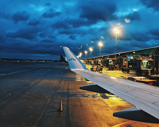 An airport runway is visible during dusk, with the wing of a KLM airplane in the foreground. Bright lights illuminate the tarmac and terminal, contrasting with the dark, cloudy sky. Various ground service vehicles and equipment are positioned near the terminal.