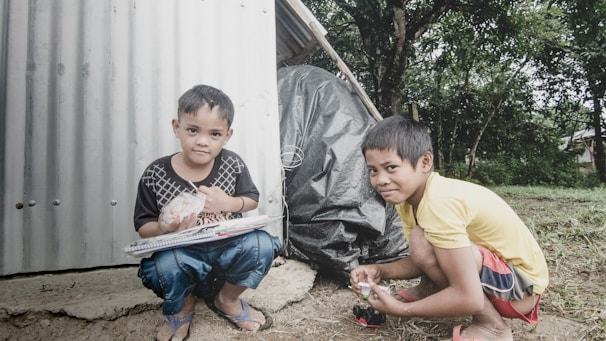 Two children are outdoors near a simple structure made of corrugated metal. One child, wearing a black shirt and jeans, is kneeling and holding a plastic-wrapped object along with a spiral notebook. The other child, wearing a yellow shirt and red shorts, is crouching and smiling, holding a small object. Trees and greenery fill the background, creating a natural setting.
