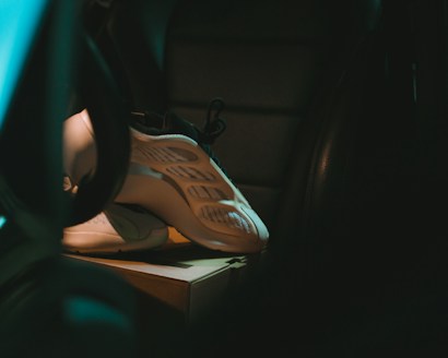 A pair of sneakers rests on a cardboard box inside a dimly lit room or vehicle interior. The shoes have a modern, sleek design and are illuminated by a soft, moody light. The setting appears enclosed, with contrasting dark shadows emphasizing the light on the sneakers.