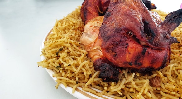 A roasted chicken is placed on a bed of spiced rice. The chicken appears to be well-cooked with a crispy, reddish-brown skin. The rice is seasoned and appears to contain small herbs and spices. This dish is presented on a white plate.