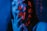 A close-up of a person with long hair bathed in blue light. Red Japanese characters are projected onto their face, creating an artistic and mysterious effect. Their hand is raised to their lips, partially obscuring their face.