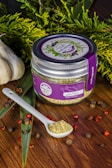 A jar labeled 'Asian Poultry' with a purple lid and label is placed on a wooden surface. The label features illustrations of herbs. A white ceramic spoon filled with a ground spice blend is positioned next to the jar. Scattered around are peppercorns and other small dried spices. Fresh greenery and whole garlic bulbs decorate the background, adding a rustic and organic touch.