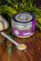 A jar labeled 'Asian Poultry' with a purple lid and label is placed on a wooden surface. The label features illustrations of herbs. A white ceramic spoon filled with a ground spice blend is positioned next to the jar. Scattered around are peppercorns and other small dried spices. Fresh greenery and whole garlic bulbs decorate the background, adding a rustic and organic touch.