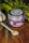 A jar labeled 'Asian Poultry' with a purple lid and label is placed on a wooden surface. The label features illustrations of herbs. A white ceramic spoon filled with a ground spice blend is positioned next to the jar. Scattered around are peppercorns and other small dried spices. Fresh greenery and whole garlic bulbs decorate the background, adding a rustic and organic touch.