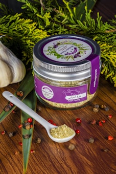 A jar labeled 'Asian Poultry' with a purple lid and label is placed on a wooden surface. The label features illustrations of herbs. A white ceramic spoon filled with a ground spice blend is positioned next to the jar. Scattered around are peppercorns and other small dried spices. Fresh greenery and whole garlic bulbs decorate the background, adding a rustic and organic touch.