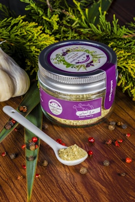 A jar labeled 'Asian Poultry' with a purple lid and label is placed on a wooden surface. The label features illustrations of herbs. A white ceramic spoon filled with a ground spice blend is positioned next to the jar. Scattered around are peppercorns and other small dried spices. Fresh greenery and whole garlic bulbs decorate the background, adding a rustic and organic touch.