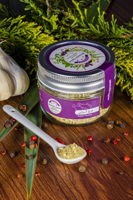 A jar labeled 'Asian Poultry' with a purple lid and label is placed on a wooden surface. The label features illustrations of herbs. A white ceramic spoon filled with a ground spice blend is positioned next to the jar. Scattered around are peppercorns and other small dried spices. Fresh greenery and whole garlic bulbs decorate the background, adding a rustic and organic touch.