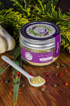 A jar labeled 'Asian Poultry' with a purple lid and label is placed on a wooden surface. The label features illustrations of herbs. A white ceramic spoon filled with a ground spice blend is positioned next to the jar. Scattered around are peppercorns and other small dried spices. Fresh greenery and whole garlic bulbs decorate the background, adding a rustic and organic touch.