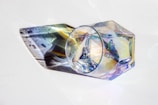 A geometric, transparent glass object with multi-faceted surfaces reflecting and refracting light. The glass creates colorful patterns and shadows against a white background, showcasing a spectrum of colors including blues, purples, and yellows.