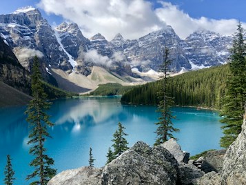 A stunning glacial lake sits surrounded by towering snow-capped mountains and lush green pine forests. The water is a vivid turquoise, reflecting the clear blue sky and fluffy white clouds above. Rocky outcroppings and vibrant green trees frame the serene landscape, adding to the natural beauty.