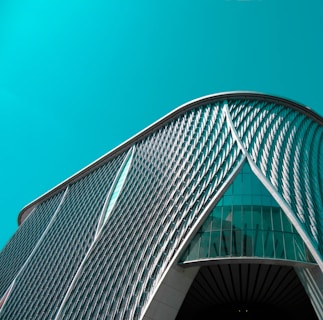 An architectural structure featuring a modern, curved design with a pattern of diagonal lines and large glass elements in the facade against a teal sky.