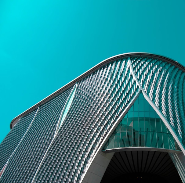 An architectural structure featuring a modern, curved design with a pattern of diagonal lines and large glass elements in the facade against a teal sky.
