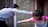 Two children are playfully having a water fight outside. One child is splashing water using a blue water balloon, while the other is preparing to throw water. The background consists of a wooden fence and blurred elements, indicating an outdoor setting.