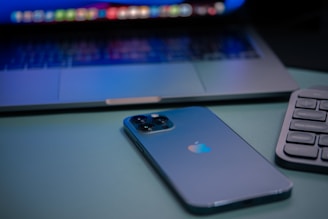 A sleek smartphone with a reflective logo is placed next to a laptop and a keyboard. The devices are arranged on a smooth surface, with the laptop partially open, displaying a colorful screen.