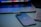 A sleek smartphone with a reflective logo is placed next to a laptop and a keyboard. The devices are arranged on a smooth surface, with the laptop partially open, displaying a colorful screen.