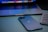 A sleek smartphone with a reflective logo is placed next to a laptop and a keyboard. The devices are arranged on a smooth surface, with the laptop partially open, displaying a colorful screen.