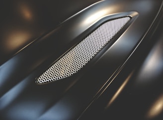 A sleek, metallic surface with a glossy finish, featuring a honeycomb mesh grille. The design is smooth and aerodynamic, suggesting it is part of a vehicle's exterior.