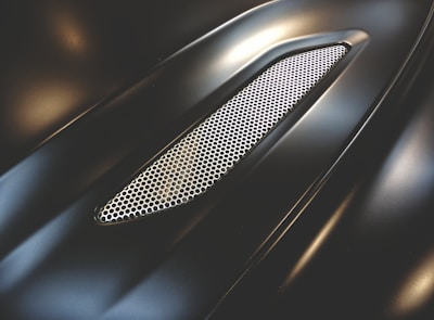 A sleek, metallic surface with a glossy finish, featuring a honeycomb mesh grille. The design is smooth and aerodynamic, suggesting it is part of a vehicle's exterior.