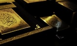 Several ornate books with intricate golden designs and Arabic calligraphy are displayed in a dimly lit setting, highlighting their elegance and detail.