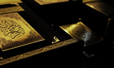 Several ornate books with intricate golden designs and Arabic calligraphy are displayed in a dimly lit setting, highlighting their elegance and detail.