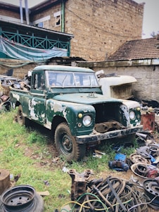 An old, green, and rusty pickup truck is parked in a yard filled with various scrap materials and car parts. The vehicle shows signs of wear, with patches of missing paint. Surrounding the truck is an assortment of metal debris and tires. The background features a stone building with a corrugated roof and exposed pipes.