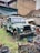 An old, green, and rusty pickup truck is parked in a yard filled with various scrap materials and car parts. The vehicle shows signs of wear, with patches of missing paint. Surrounding the truck is an assortment of metal debris and tires. The background features a stone building with a corrugated roof and exposed pipes.