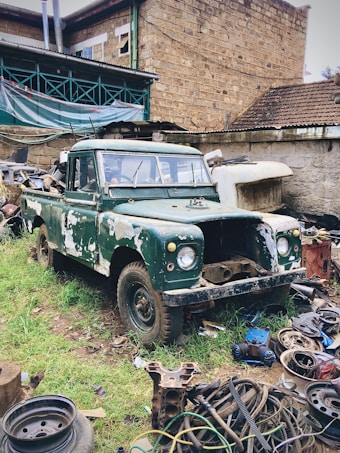 An old, green, and rusty pickup truck is parked in a yard filled with various scrap materials and car parts. The vehicle shows signs of wear, with patches of missing paint. Surrounding the truck is an assortment of metal debris and tires. The background features a stone building with a corrugated roof and exposed pipes.