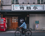 A street scene featuring a storefront with a large sign in Japanese characters. A red Coca-Cola vending machine stands on the sidewalk beside wooden doors. In front of the closed doors is a person riding a bicycle, bundled in a blue jacket and helmet. The storefront has traditional Japanese elements, including a fabric curtain with a logo. A chalkboard on an easel is placed near a rolling shutter.