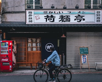 A street scene featuring a storefront with a large sign in Japanese characters. A red Coca-Cola vending machine stands on the sidewalk beside wooden doors. In front of the closed doors is a person riding a bicycle, bundled in a blue jacket and helmet. The storefront has traditional Japanese elements, including a fabric curtain with a logo. A chalkboard on an easel is placed near a rolling shutter.