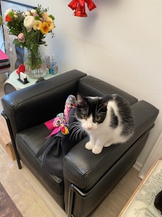 A black and white cat sits on a black leather armchair, next to a stylish black handbag adorned with a colorful scarf. In the background, there is a bouquet of vibrant flowers in a glass vase and some decorative items on a small table against a plain white wall.