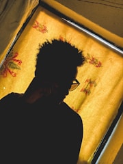 A silhouette of a person with curly hair and glasses, seated in front of a window with a yellow curtain adorned with floral patterns.