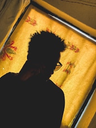 A silhouette of a person with curly hair and glasses, seated in front of a window with a yellow curtain adorned with floral patterns.