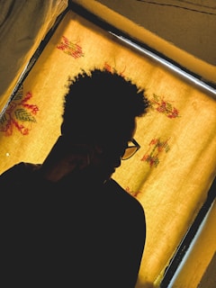 A silhouette of a person with curly hair and glasses, seated in front of a window with a yellow curtain adorned with floral patterns.