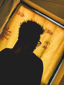 A silhouette of a person with curly hair and glasses, seated in front of a window with a yellow curtain adorned with floral patterns.