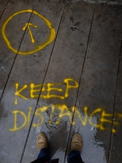 Yellow spray-painted text on a wet concrete surface reads 'KEEP DISTANCE'. A yellow circle with an upward arrow is on the left. Two brown shoes are visible at the bottom of the image, hinting at a person standing on the patchy, reflective surface. Raindrop patterns or water stains are apparent on the ground.