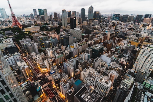 A sprawling cityscape filled with densely packed skyscrapers and buildings, stretching into the distance under a cloudy sky. Bright city lights illuminate the streets, creating a vibrant and bustling atmosphere. A prominent red tower stands out among the grey and glass structures.
