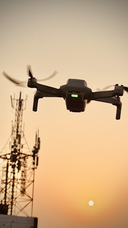 A drone is flying in the foreground, captured against a backdrop of a sunset. The sky is golden and clear, providing a striking contrast to the dark silhouette of a communication tower on the left side of the image.