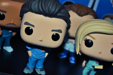Vinyl figurines with oversized heads and cartoonish features dressed in blue clothing, standing on a dark surface. The figures exhibit exaggerated expressions and styles, hinting at character portrayals. Additional figures can be seen partially in the background.