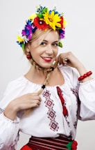 A woman with blonde hair styled in a braid wears a traditional embroidered white blouse. She accessorizes with a vibrant floral headpiece consisting of colorful flowers such as yellow, red, blue, and purple. Red beads adorn her wrists and belt. She exudes a cheerful and confident expression.