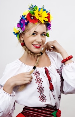 A woman with blonde hair styled in a braid wears a traditional embroidered white blouse. She accessorizes with a vibrant floral headpiece consisting of colorful flowers such as yellow, red, blue, and purple. Red beads adorn her wrists and belt. She exudes a cheerful and confident expression.