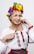A woman with blonde hair styled in a braid wears a traditional embroidered white blouse. She accessorizes with a vibrant floral headpiece consisting of colorful flowers such as yellow, red, blue, and purple. Red beads adorn her wrists and belt. She exudes a cheerful and confident expression.