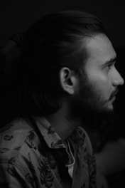 A black and white profile portrait of a man with long hair tied back. He has a beard and is wearing a patterned shirt. The lighting is soft, highlighting the contours of his face against a dark background.