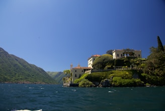 A picturesque lakeside villa is perched on a lush, green hillside. The architecture features pale yellow walls and terracotta roofs, surrounded by manicured gardens and tall trees. The lake extends towards the mountains in the background under a clear blue sky.