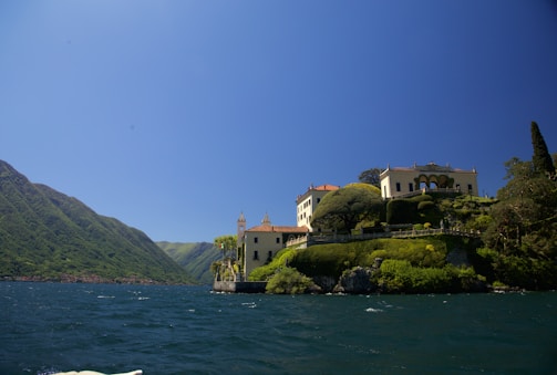 A picturesque lakeside villa is perched on a lush, green hillside. The architecture features pale yellow walls and terracotta roofs, surrounded by manicured gardens and tall trees. The lake extends towards the mountains in the background under a clear blue sky.