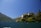 A picturesque lakeside villa is perched on a lush, green hillside. The architecture features pale yellow walls and terracotta roofs, surrounded by manicured gardens and tall trees. The lake extends towards the mountains in the background under a clear blue sky.