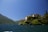 A picturesque lakeside villa is perched on a lush, green hillside. The architecture features pale yellow walls and terracotta roofs, surrounded by manicured gardens and tall trees. The lake extends towards the mountains in the background under a clear blue sky.