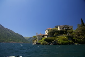 A picturesque lakeside villa is perched on a lush, green hillside. The architecture features pale yellow walls and terracotta roofs, surrounded by manicured gardens and tall trees. The lake extends towards the mountains in the background under a clear blue sky.