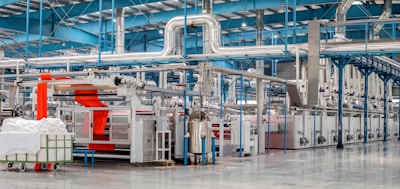 A spacious industrial facility with metal machinery and equipment, featuring a production line with a focus on textile processing. Brightly colored fabrics are moving through the machines, and there are large rolls of fabric and textile materials nearby. The environment is clean and organized, with a high ceiling and visible ductwork.