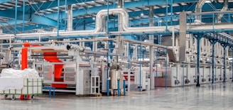 A spacious industrial facility with metal machinery and equipment, featuring a production line with a focus on textile processing. Brightly colored fabrics are moving through the machines, and there are large rolls of fabric and textile materials nearby. The environment is clean and organized, with a high ceiling and visible ductwork.